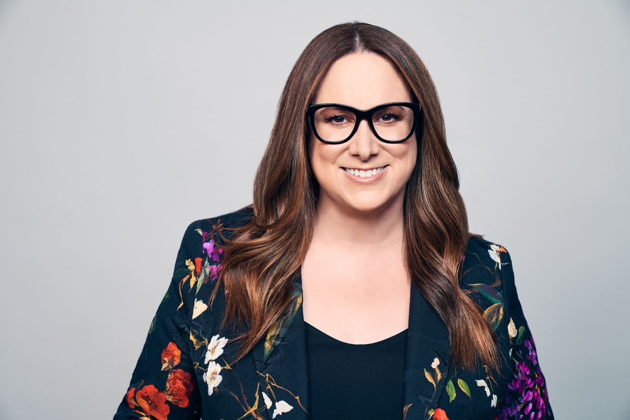 Confident woman with glasses in a floral blazer smiles warmly.